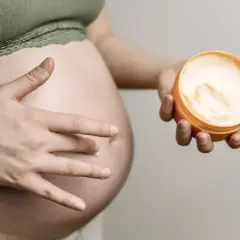 Ciało w ciąży, dłoń dotyka brzucha, druga trzyma krem. Delikatna pielęgnacja, może szczotkowanie na sucho w ciąży?