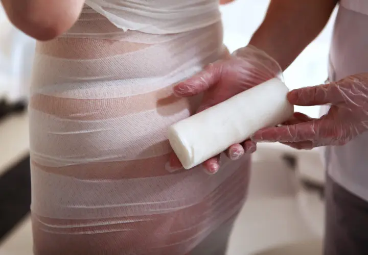 Profesjonalne body wrapping kosmetyki w trakcie zabiegu. Ręce w rękawiczkach trzymają rolkę bandaża.