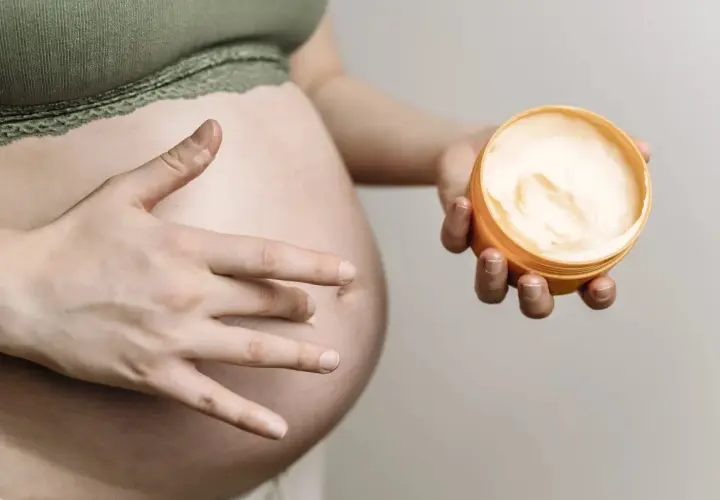 Ciało w ciąży, dłoń dotyka brzucha, druga trzyma krem. Delikatna pielęgnacja, może szczotkowanie na sucho w ciąży?