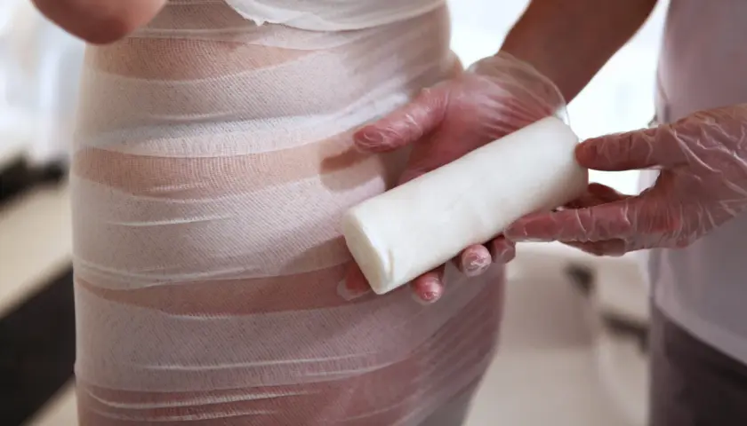 Profesjonalne body wrapping kosmetyki w trakcie zabiegu. Ręce w rękawiczkach trzymają rolkę bandaża.