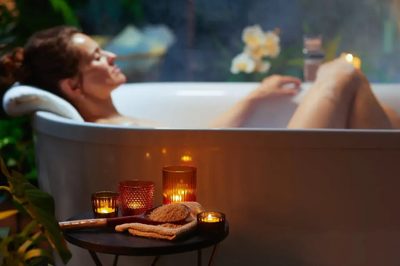 Relaks w wannie z solą do kąpieli. Kobieta odpoczywa w otoczeniu świec i zieleni, czując, co daje sól do kąpieli: ukojenie i odprężenie.