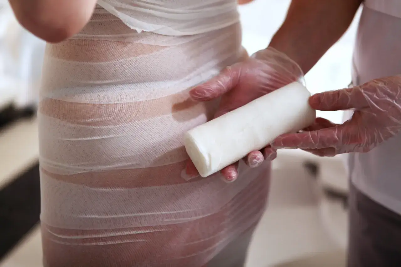 Profesjonalne body wrapping kosmetyki w trakcie zabiegu. Ręce w rękawiczkach trzymają rolkę bandaża.